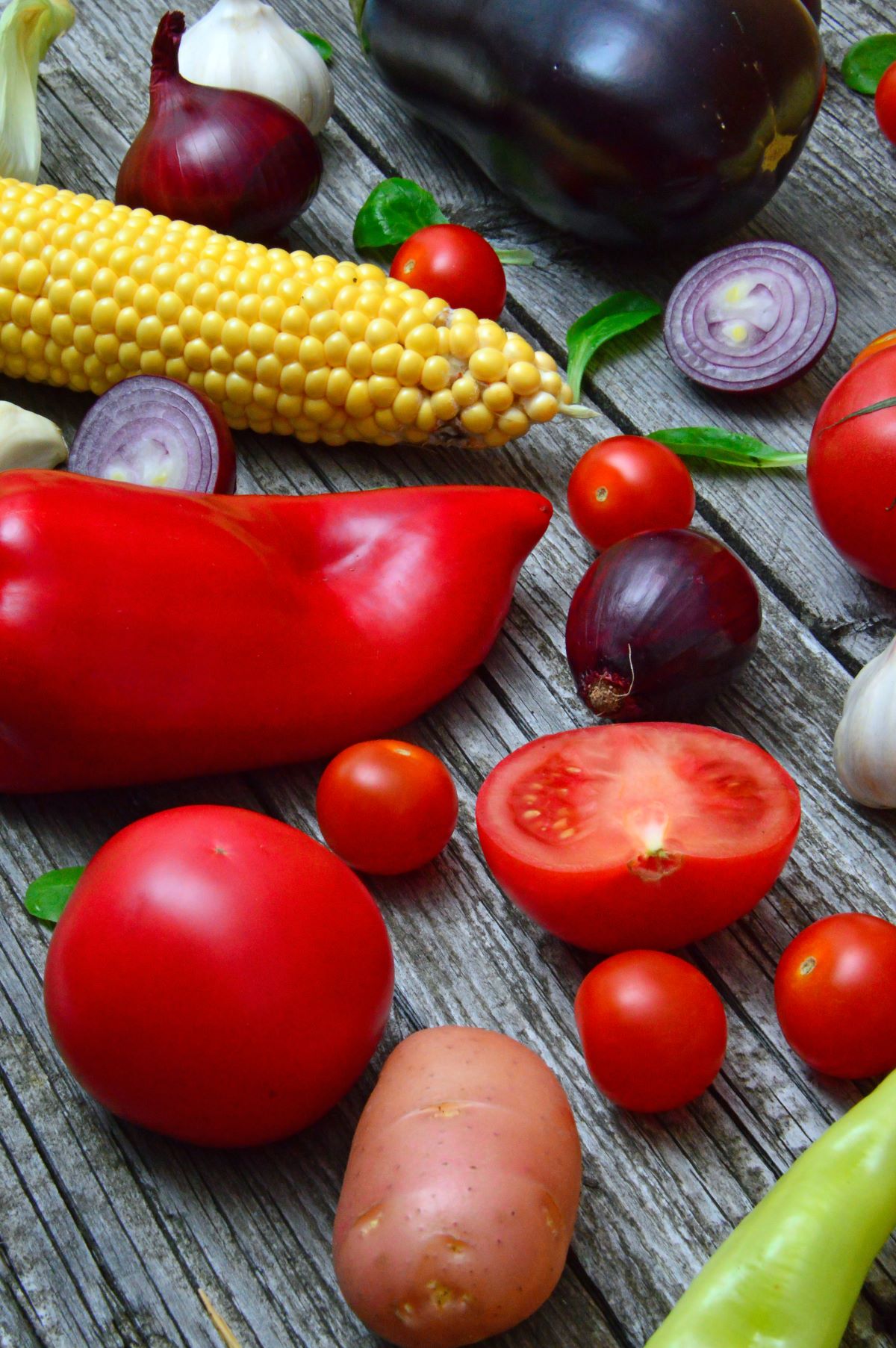 COLORFUL VEGETABLES SITTING ON A WOODEN TABLE INCLUDING, LARGE RED PEPPERS, RED ONION, TOMATOES, POTATO, GARLIC, CORN, BASIL LEAVES, AND EGGPLANT