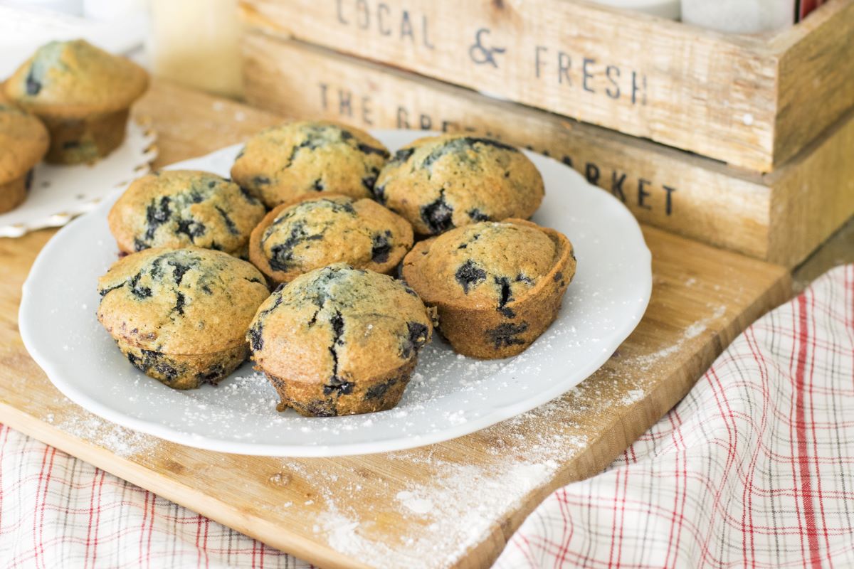 Muffin of the Month Clubs - Blueberry muffins on a white plate dusted in four sitting on a wooden cutting board in front of a wooden crate that says Local & Fresh,  and a red and gray lined napkin under the cutting board