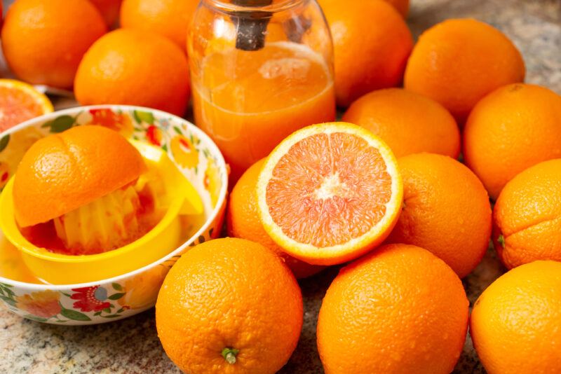 a spread of fresh cara cara oranges with a brightly printed bowl with a juicer and half orange, a glass pitcher at the middle with orange juice