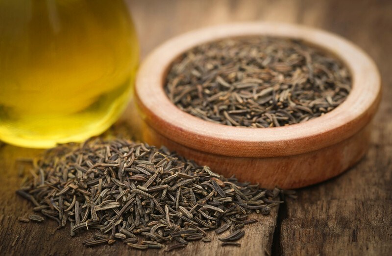 wooden bowl full of caraway seeds on a wooden surface, with a mound of caraway seeds at the front and glass of oil at the side