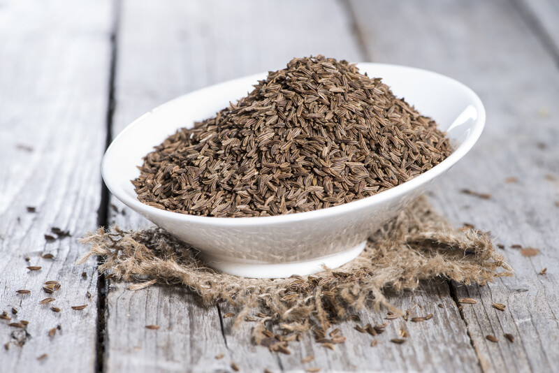 Elegant asymmetrical white dish full of caraway seeds on a small piece of brown sack, resting on an old wooden table with loose caraway seeds around it.