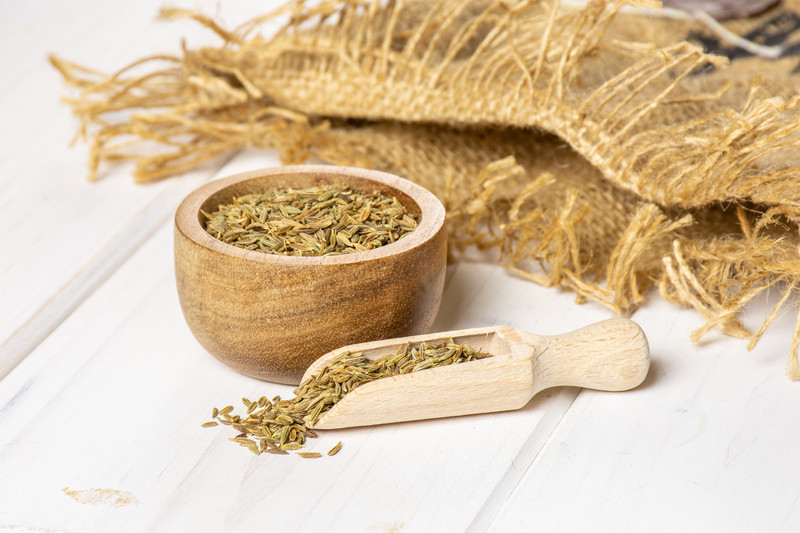 Wooden bowl and scoop full of caraway, resting on top of white wooden surface with a piece of sack cloth at the back.