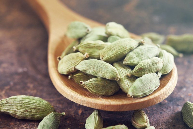 closeup image of a wooden ladle full of cardamom pods on a rusty surface with loose cardamom pods around it