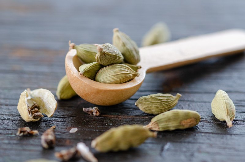 Closeup image of a small wooden scoop full of cardamom pods, with scattered cardamom pods around it.