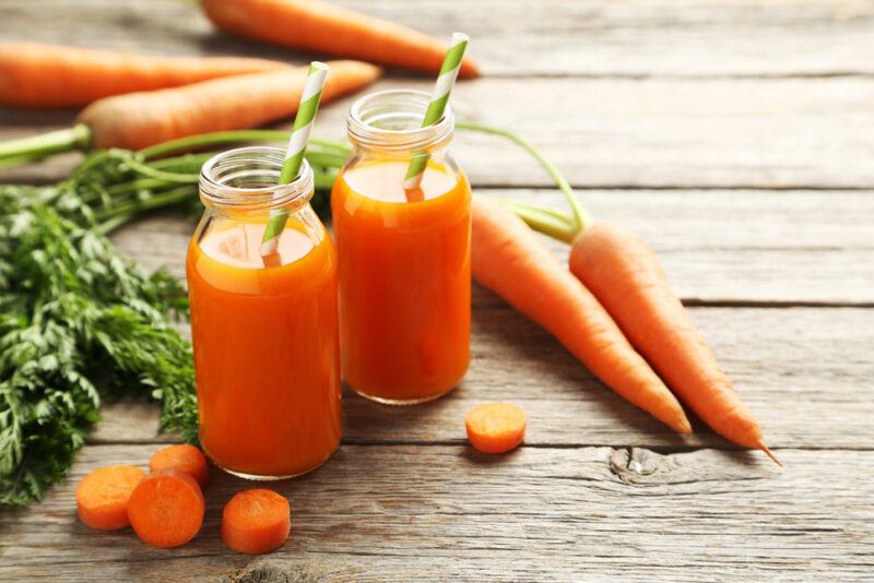 Two open jars of carrot juice with green and white straws in them rest on a wooden surface near a few carrot slices and a few whole carrots.