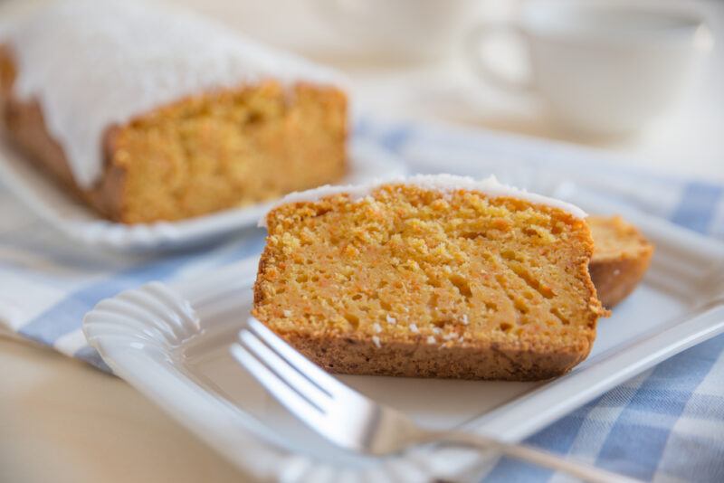 a slice of carrot cake on a white ceramic dish with a fork beside it, at the back is the rest of the carrot cake with white frosting, both are resting on top of a white table napkin with blue checkered stripes