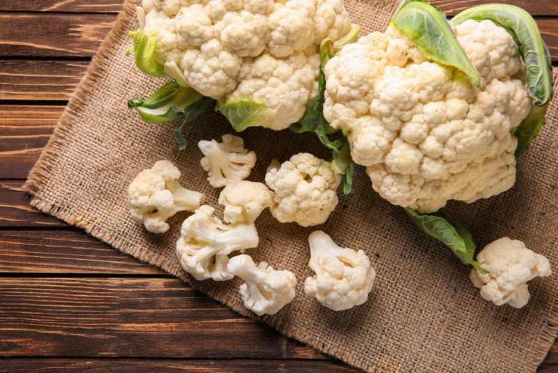 Raw cauliflower florets and a head of a cauliflower on a sack and a wooden table