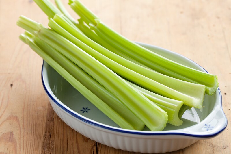 This photo shows a silver metal bowl filled with green celery stalks on a wooden table.