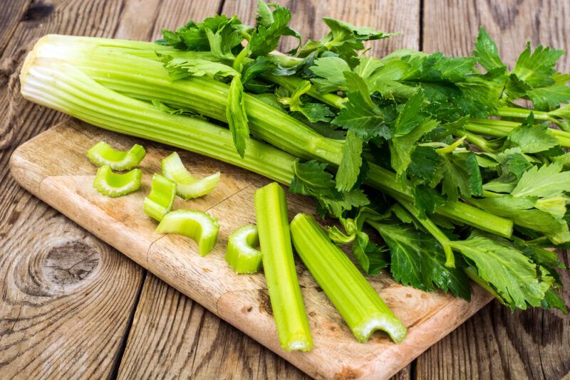 A bunch of green celery, two celery sticks, and a few celery slices rest on a wooden cutting board on a rustic wooden surface.