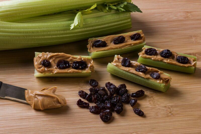 A wooden table with a snack called ants on a log, which is made using celery, peanut butter, and raisins, which are on the table as well