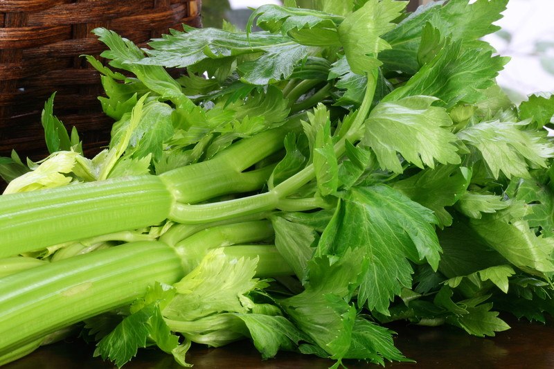 A bundle of light green celery lies on a dark tabletop next to a brown wicker basket.