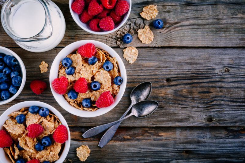 Two bowls of bran cereal on a wooden table, with milk and some fruit