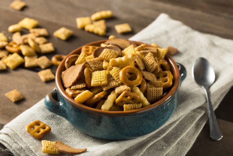 A bowl of cereal mix and a spoon rest on a beige cloth on a wooden table near some loose pieces of the mix.