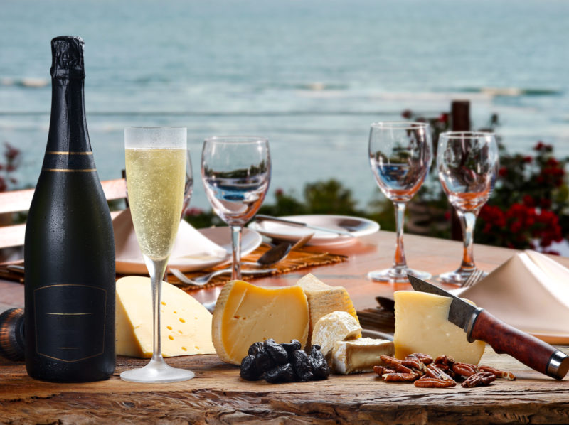 A cheese board set up outside overlooking the ocean, including wine glasses, a bottle of champagne and a flute of champagne