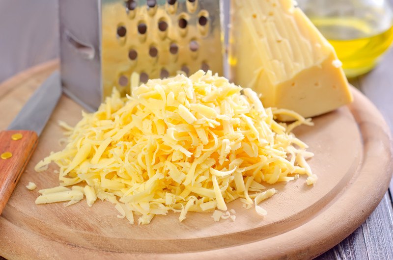 This photo shows a cheese grater, a block of cheese, and some shredded cheese on a cutting board that’s resting on a wooden table.