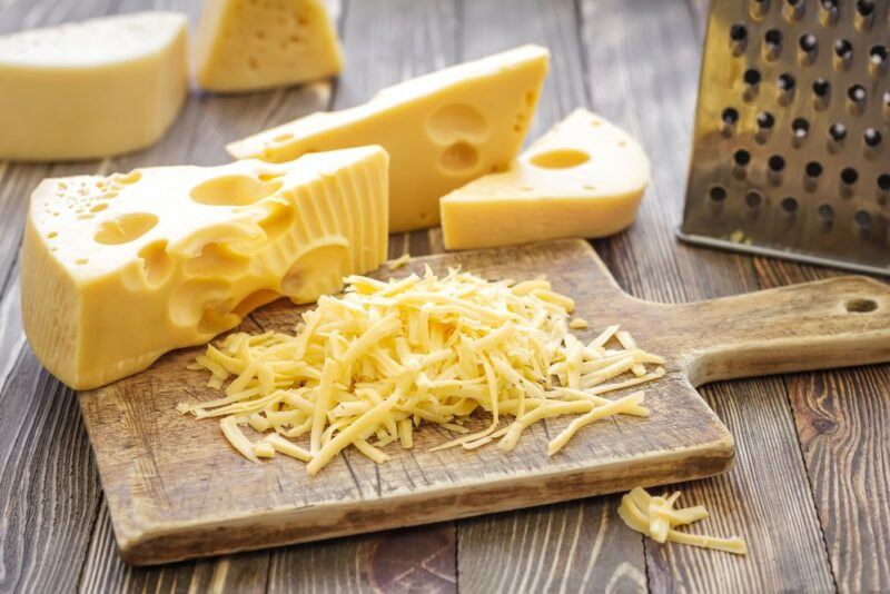 A wooden board with grated cheese, in front of some blocks of cheese and a grater