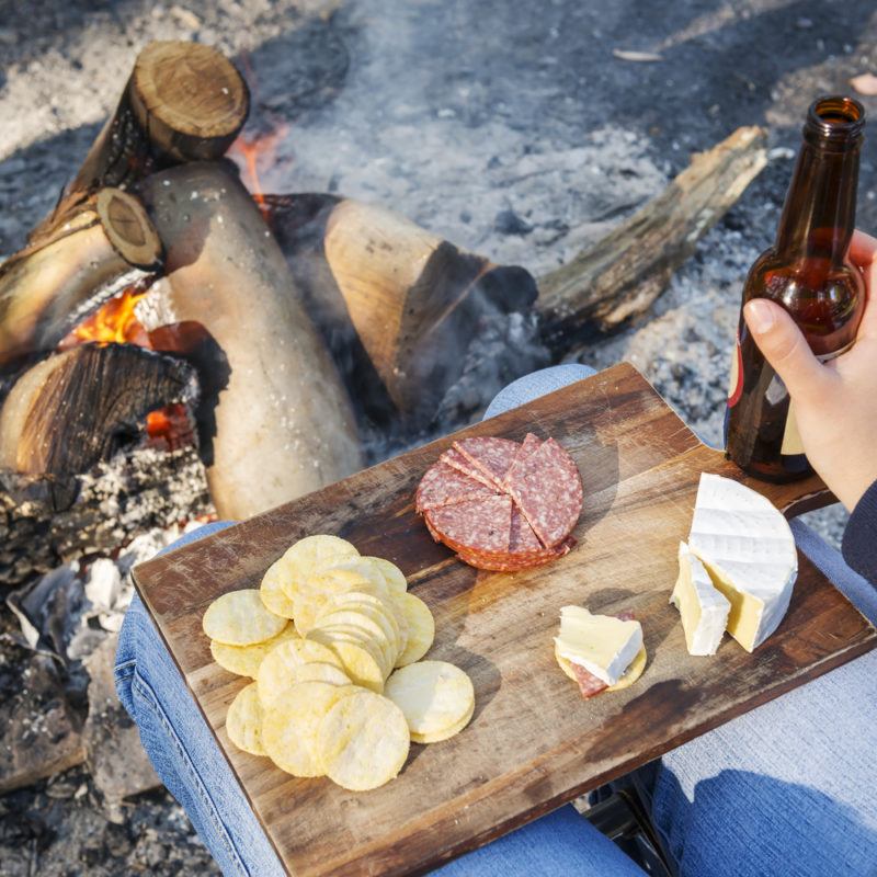 A wooden board with cheese, meat, and crisps near a campfire