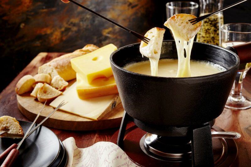 A black fondue pot with cheese, where bread is being dipped into it, next to a wooden board containing cheese