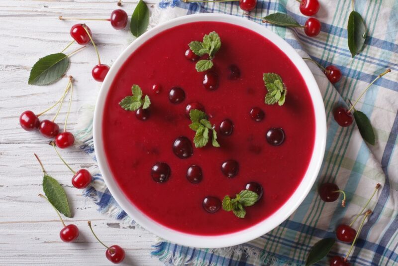 A large white bowl containing cherry soup with green leaves, with more cherries and leaves scattered on the table