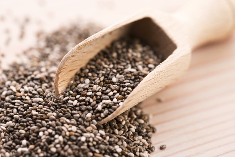 This photo shows a wooden scoop of chia seeds spilling onto a wooden table.