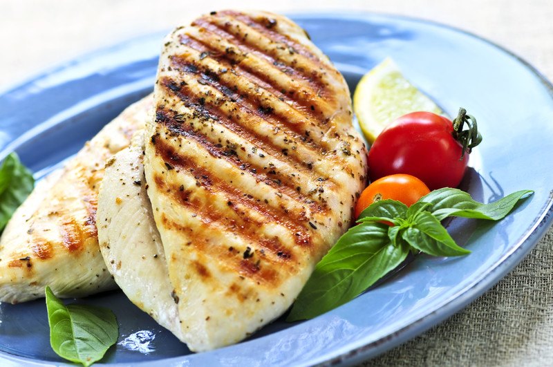 Several pieces of grilled chicken rest on a blue plate with tomatoes, a lemon wedge, and mint leaves.