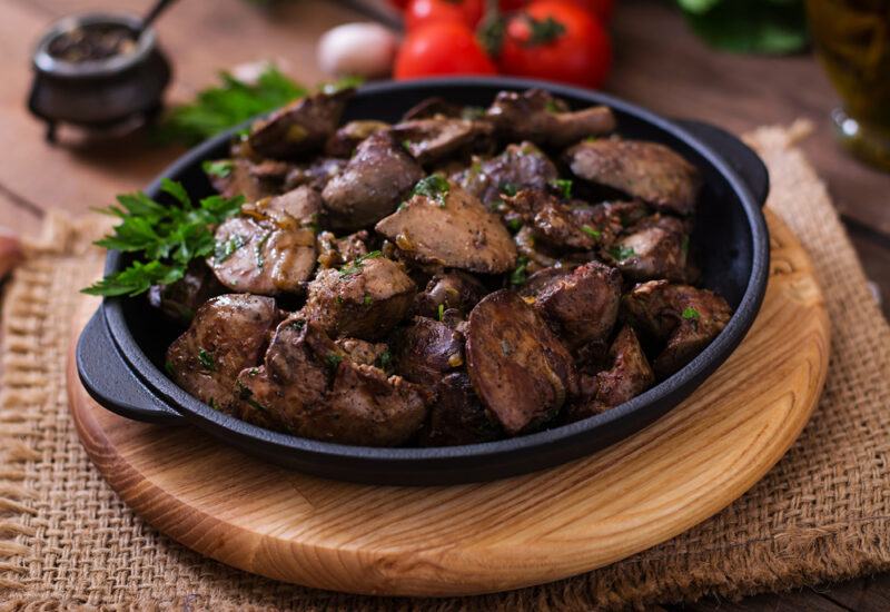 Fried,Chicken,Liver,With,Onions,And,Herbs | Food For Net This photo shows a dark plate of cooked chicken liver on a piece of wood and burlap, in front of a background containing tomatoes.
