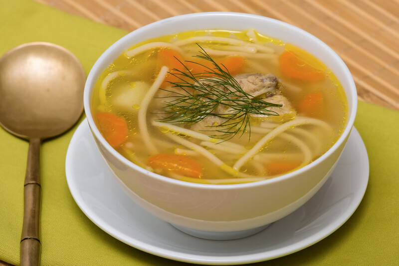 This photo shows a white bowl filled with chicken soup, with a spoon lying on a green cloth on the left side of the bowl.