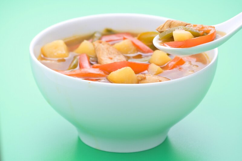 A white ceramic bowl filled with chicken soup and a white ceramic spoon with soup in it rest against a green background.