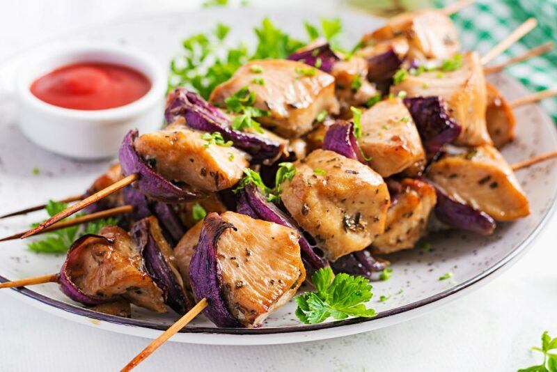 A white plate with chicken and red onion kebabs and a small pot of dip in the background