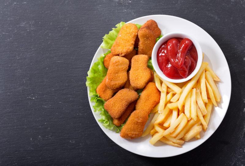 A white plate with processed chicken nuggets, tomato sauce, and fries