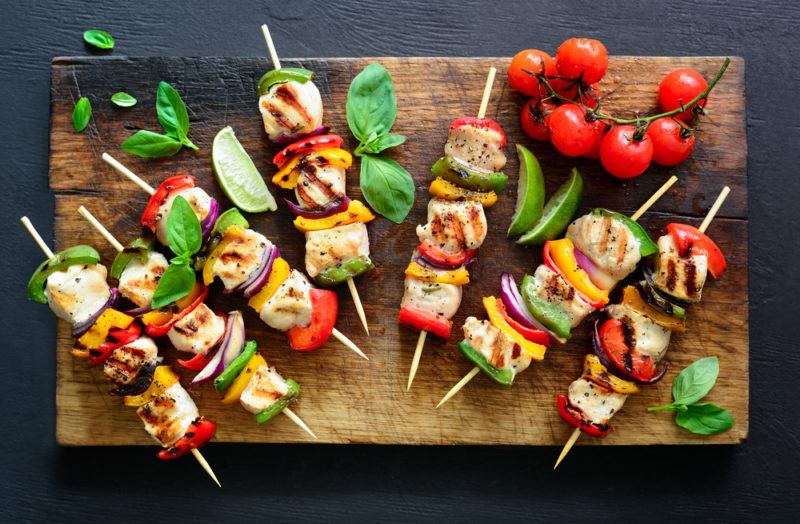 Brightly colored skewers that inlcude chicken and cooked peppers, on a cutting board with leaves and tomatoes