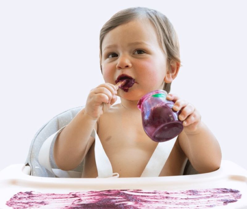 Baby sitting in high chair eating a purple baby food puree - baby food smeared all over the table top of the high chair 