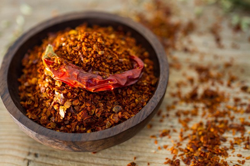 a wooden bowl full of chili pepper flakes with dried pepper on top and lose chili pepper flakes around it