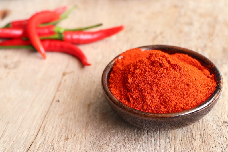 Brown ceramic bowl full of chili powder on a wooden surface with blurred image of fresh chili peppers at the back.