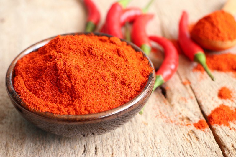 brown ceramic bowl full of chili powder on a wooden surface with fresh chilis at the back and wooden spoon full of chili powder