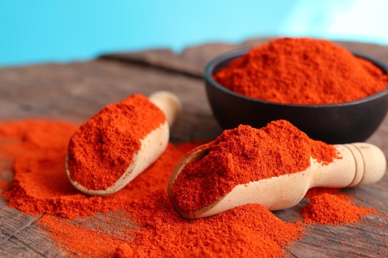 a couple of wooden scoops full of chili powder and loose chili powder around it on a wooden table, with a black bowl full of chili powder at the back