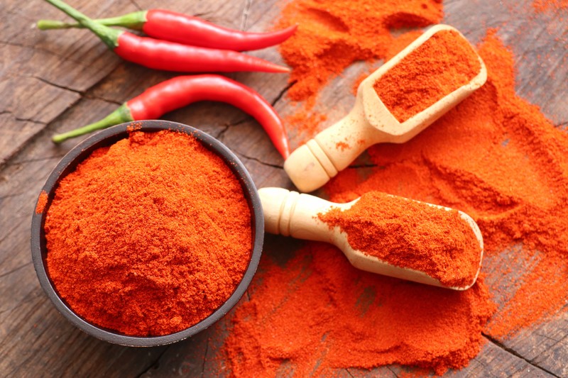 top view image of a bowl and two wooden scoops full of chili powder, on a wooden table with fresh chilis