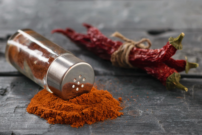 a small mound of chili powder on a stone surface with empty small spice jar toppled over on top of it and dried chili tied with a string at the back