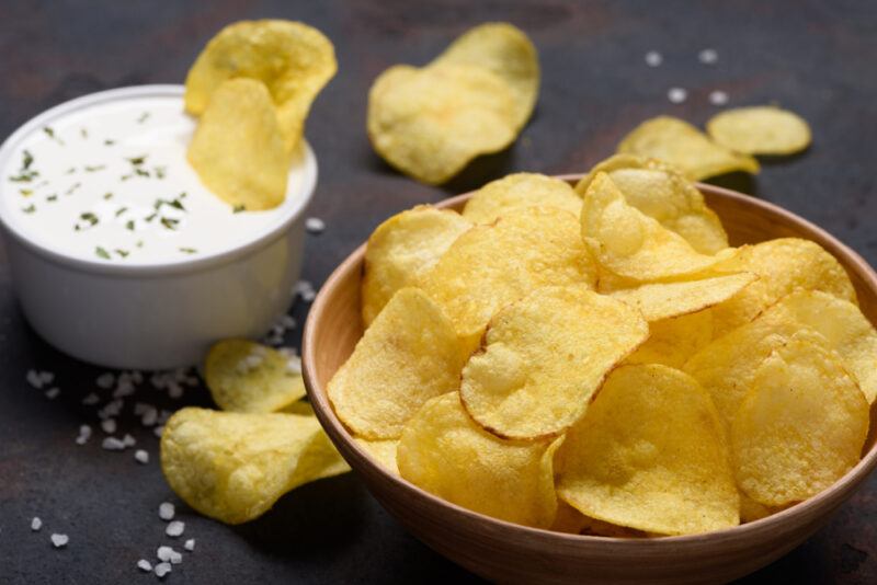 A bowl of yellow potato chips rests on a dark surface near a white tub of white dip with green seasonings.