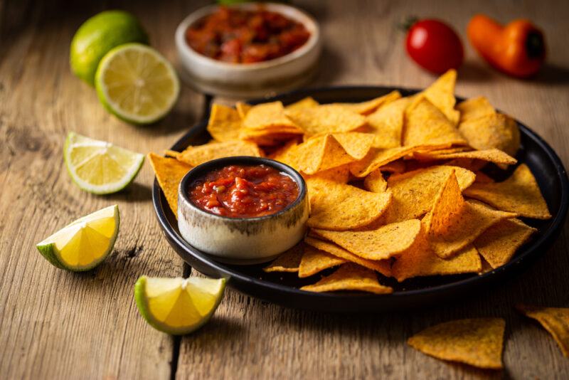 A dark plate of tortilla chips with a small dish of salsa rests on a rustic wooden surface near some lime slices, more salsa, a cherry tomato, and an orange pepper.