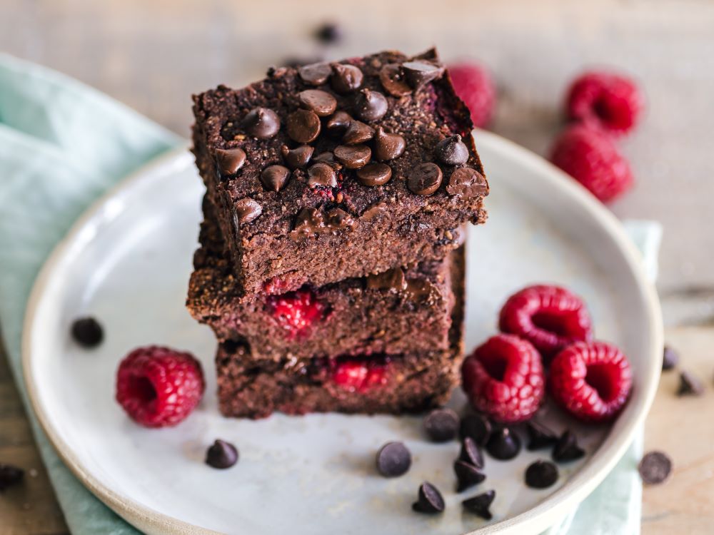 Brownie of the Month Club, 3 double chocolate raspberry brownies stacked on a plate with raspberries and chocolate chips artfully placed around the white plate and napkin 