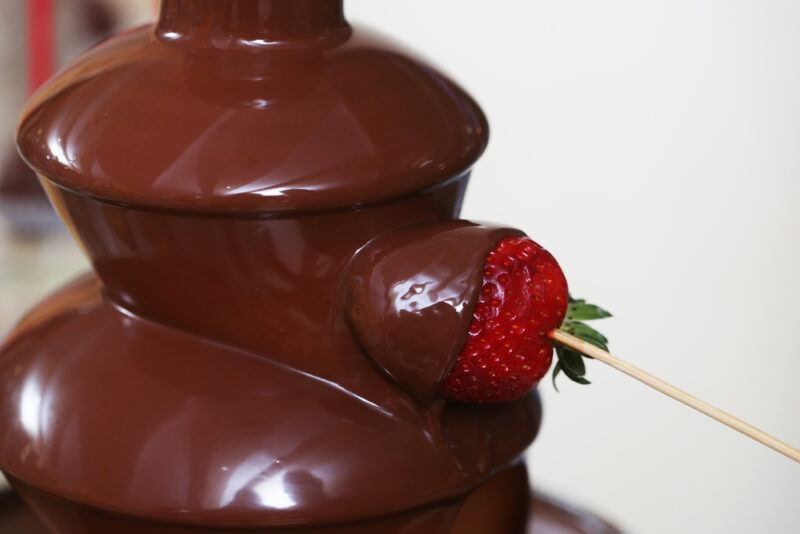 A strawberry on a wooden kabob stick is being dipped into a chocolate fountain.