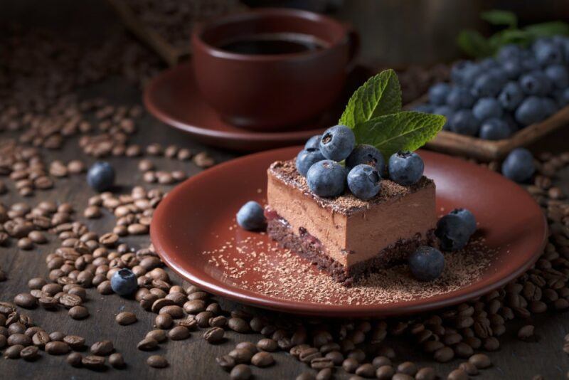 A brown plate with chocolate cake or a chocolate cheesecake that's been topped with blueberries. There are coffee beans on the table.