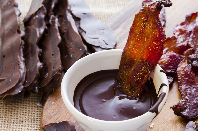 A stripe of bacon dipped in chocolate besides chocolate covered bacon.