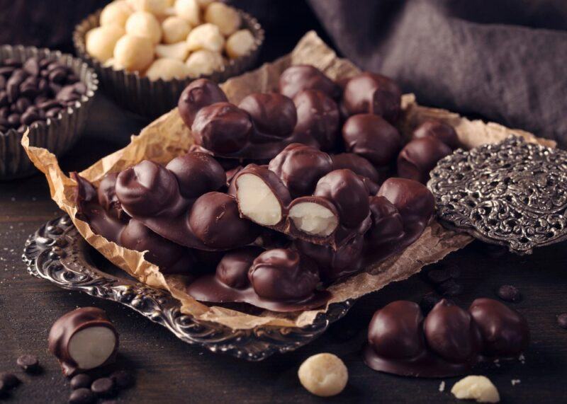 A dark brown dish containing chocolate covered macadamia nuts, with a small bowl of macadamia nuts in the background