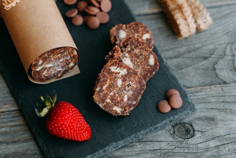 A black slate block with a roll of chocolate salami, next to some chocolate pieces and a strawberry