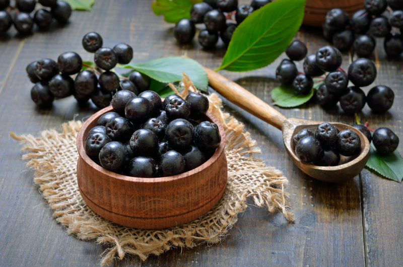 A wooden bowl filled with chokeberries, next to a spoon of the berries and various berries scattered across a wooden table