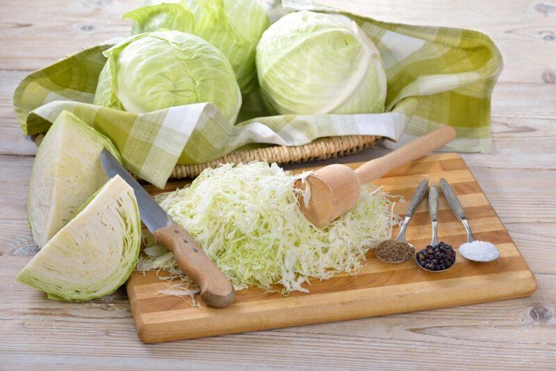 A woden board with chopped cabbage for making sauerkraut, with more whole and partial cabbages in the background