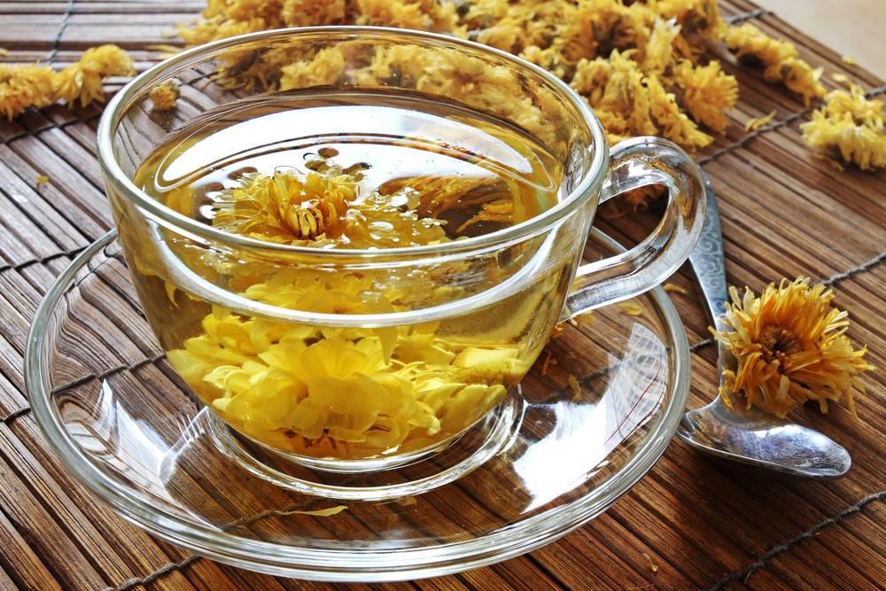 A glass mug of clear tea with chrsanthemum flowers, with more flowers scattered on the wooden table