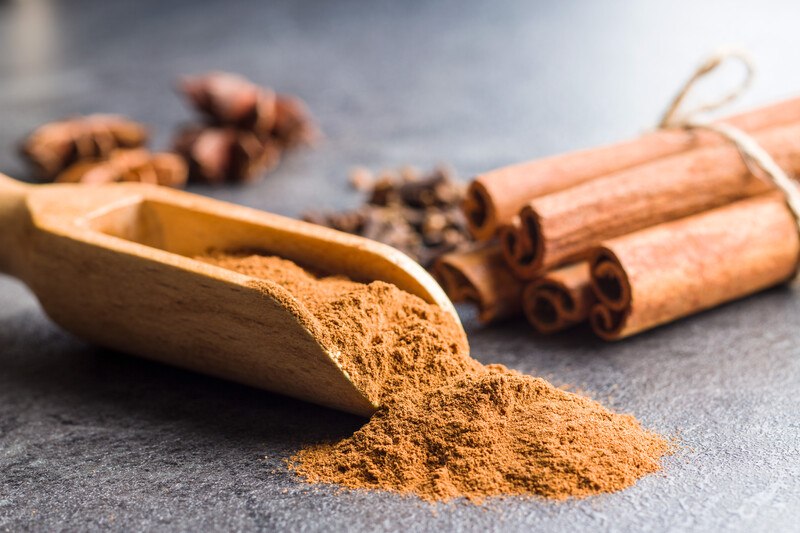 a wooden scoop full of ground cinnamon on a black surface, with cinnamon sticks tied together with a string at the back